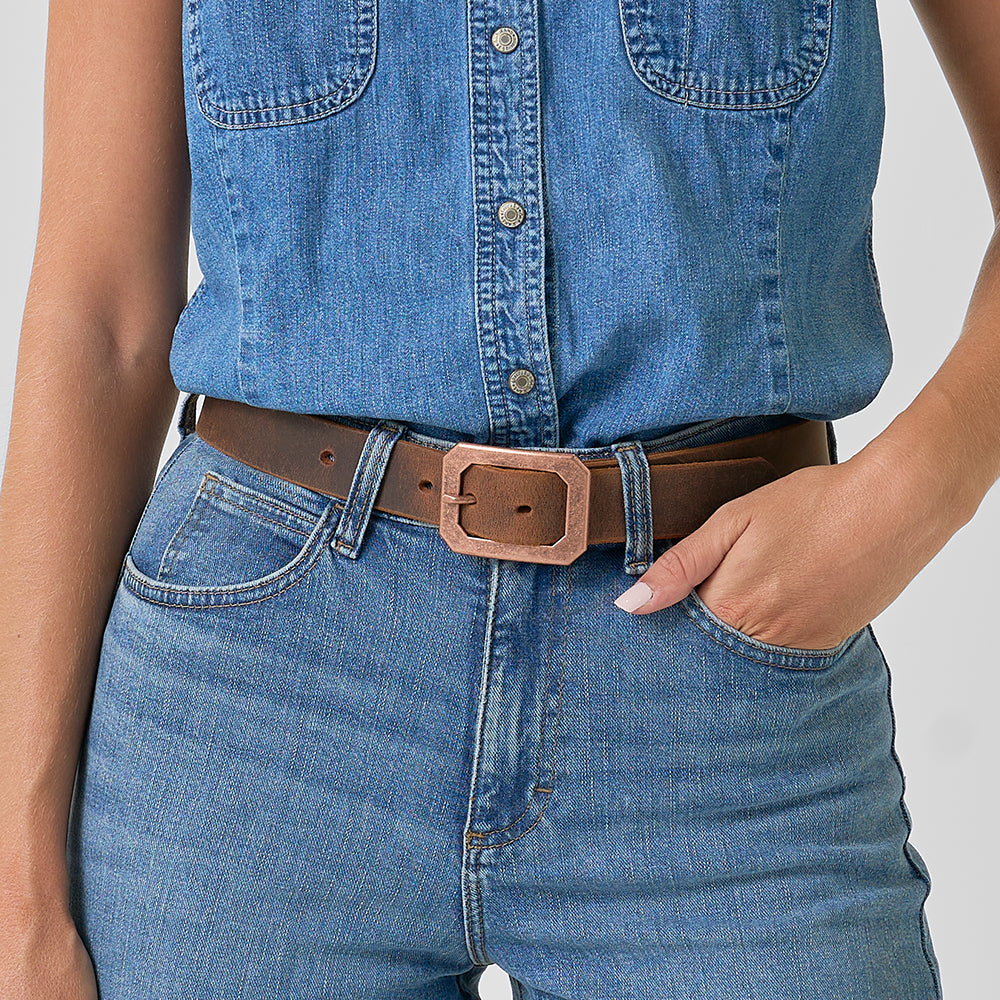 Wearing a sleeveless denim shirt and high-waisted blue jeans, this person styles their look with The Lainey by Hanks Belts—a western-inspired brown leather belt with a rectangular buckle—and poses with one hand in their pocket.