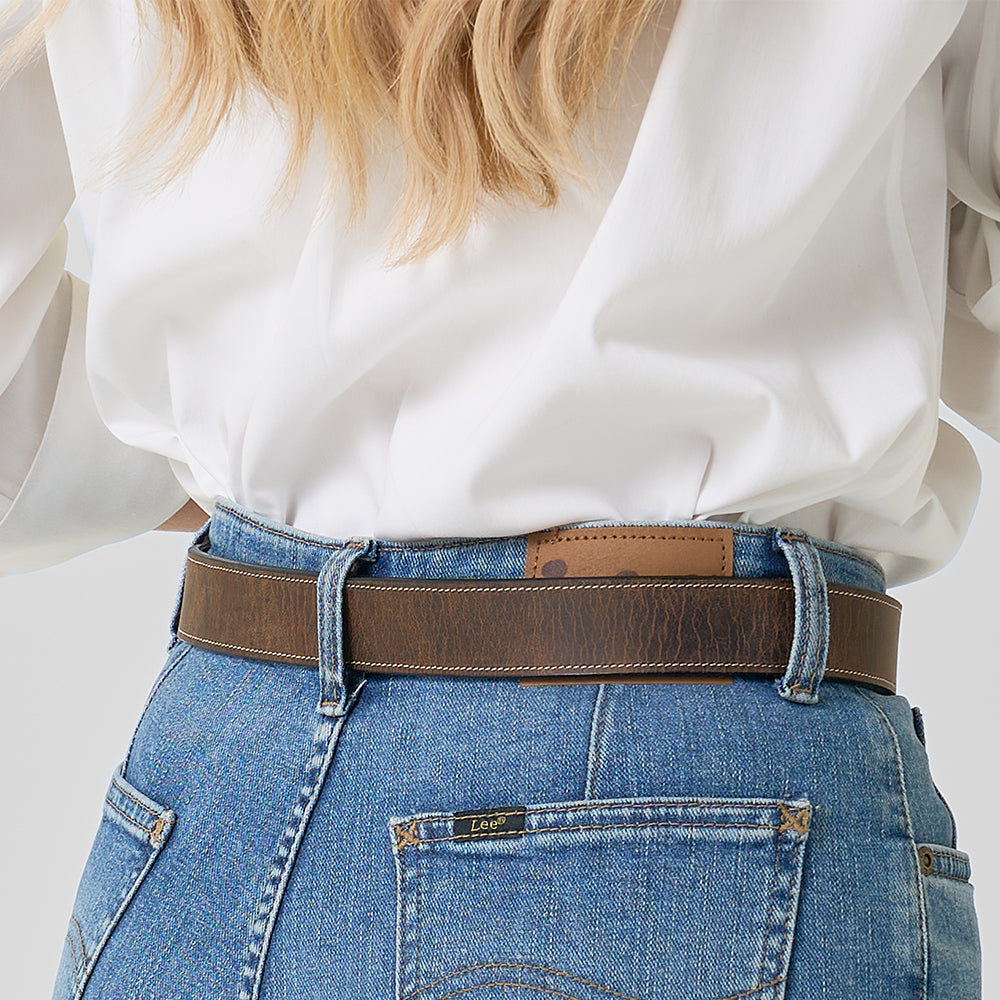 A person in blue jeans with a "Lee" label wears Hanks Belts' The Willow Bison, a brown leather western belt, over a tucked-in white shirt; long, wavy blonde hair is partially visible.