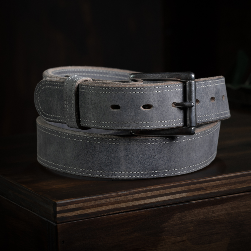 The Montana Bison Double Layer by Hanks Belts, featuring rugged bison leather, white stitching, and a matte black buckle, is coiled neatly on a dark wooden surface.