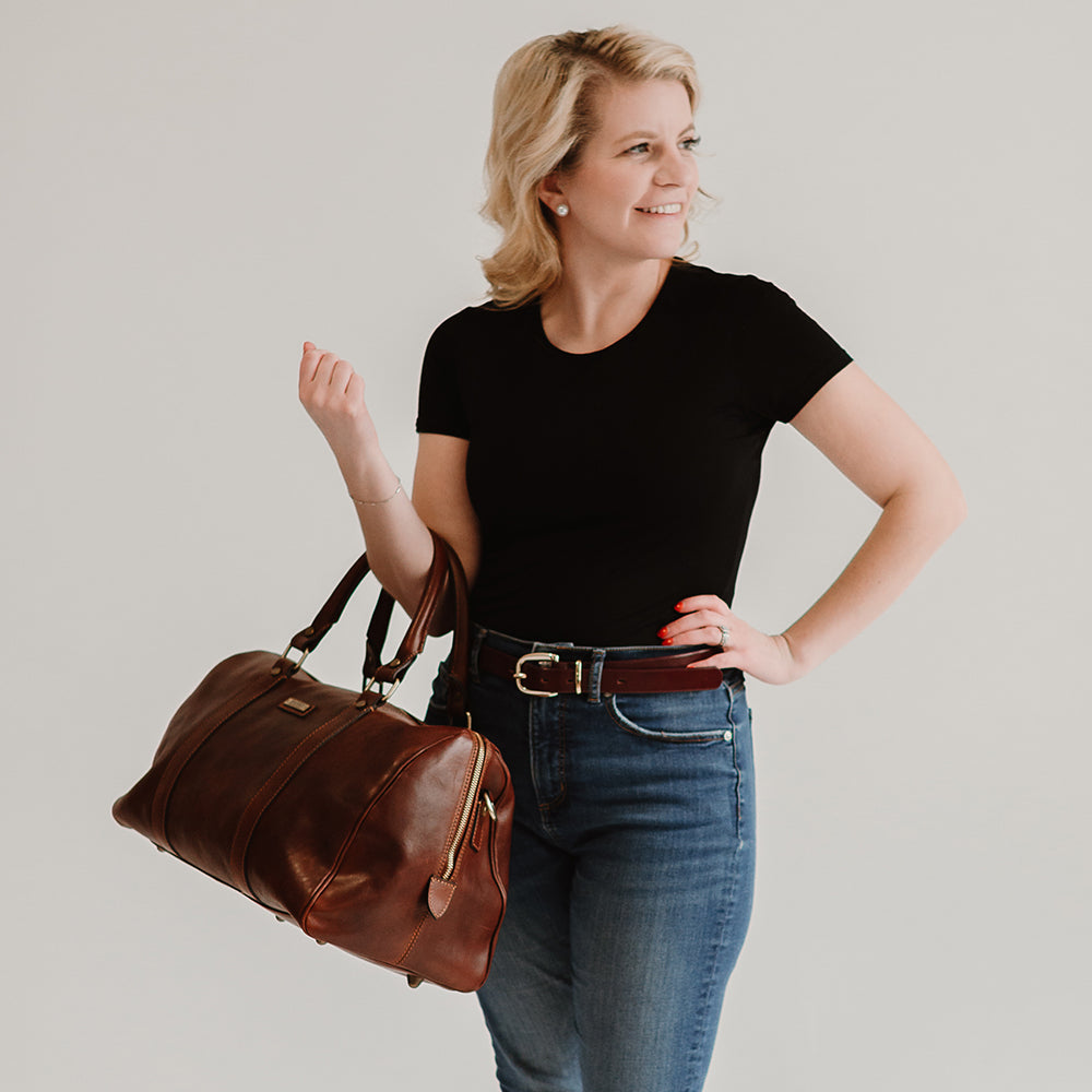 A blonde woman in a black t-shirt and jeans holds the Hanks Belts' Getaway Duffel With Luggage Sleeve, made from full-grain Italian leather. She stands against a light background, smiling and looking to the side, exemplifying an ideal travel companion.