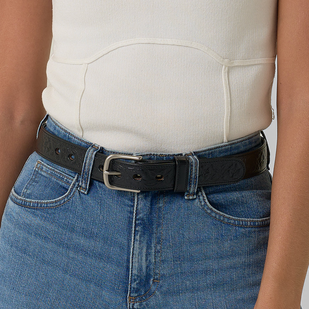 A person wears a cream sleeveless top tucked into blue jeans, accessorized with The Floral Embossed women's leather belt from Hanks Belts featuring a silver buckle.