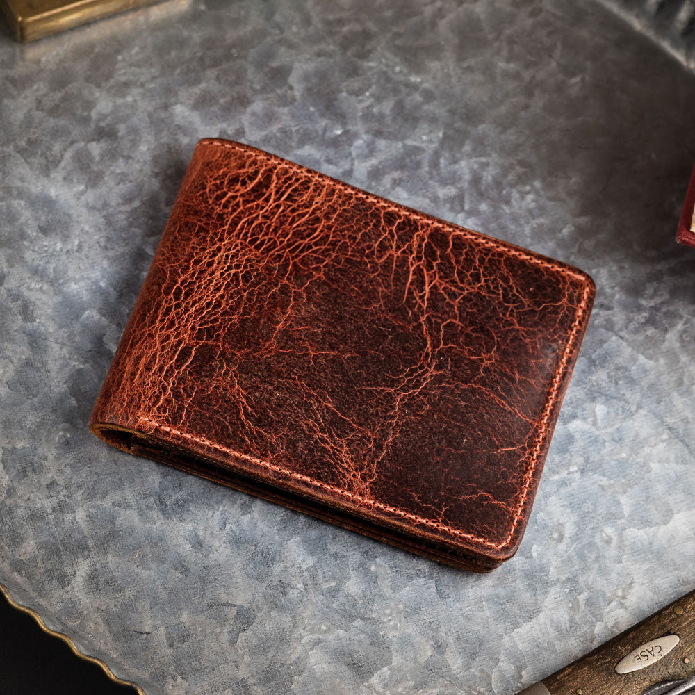 The Lanier Premium Bison Bifold by Hanks Belts rests closed on a textured gray metal surface, highlighting its rugged brown distressed bison leather and distinctive USA-made exterior.