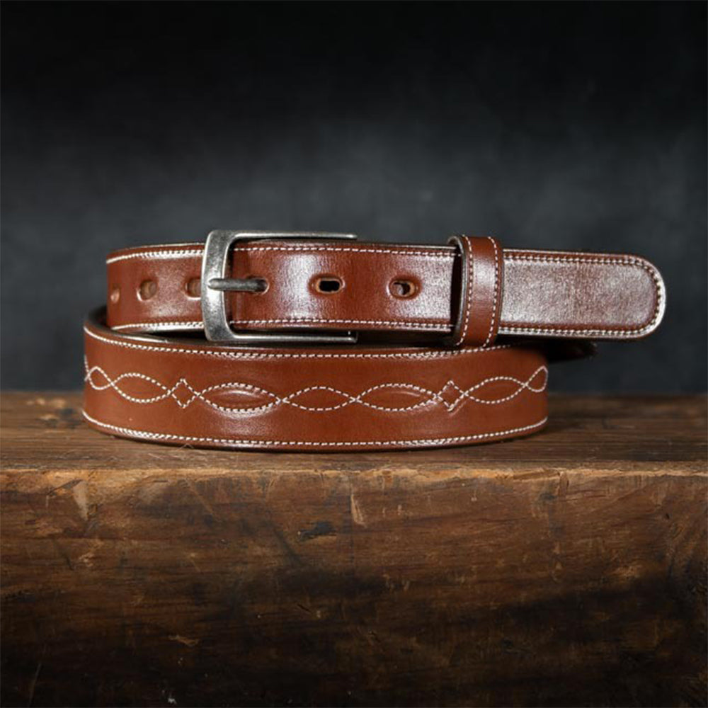 The Teton Tapered by Hanks Belts, made from vegetable tanned leather with white infinity stitching and a silver buckle, rests coiled on a rustic wooden surface against a dark background.