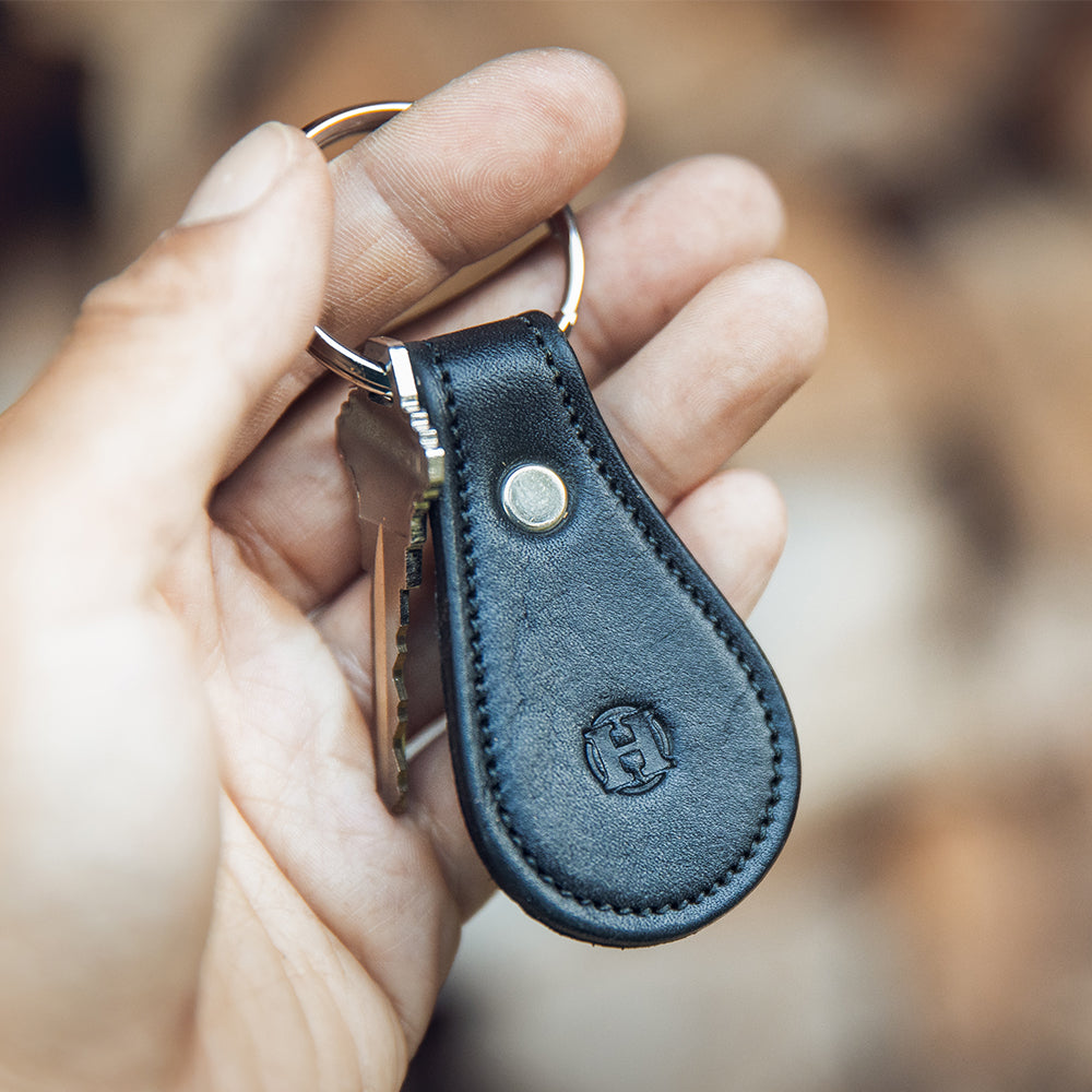 In a softly blurred background emphasizing its elegant craftsmanship, a hand holds the Black Bell-Shaped Leather Keychain from Hanks. Crafted from full grain leather, it features a metal rivet and an embossed "H" logo, with a single key attached to the keyring.