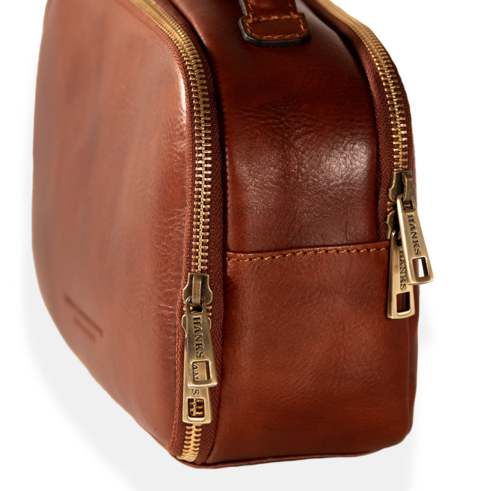 Close-up of a luxurious Tidy Travel Case by Hanks Belts, featuring two brass zippers engraved with the brand's name. Crafted with Italian craftsmanship, the smooth leather surface showcases visible stitching against a plain white background, making it an ideal toiletry bag.