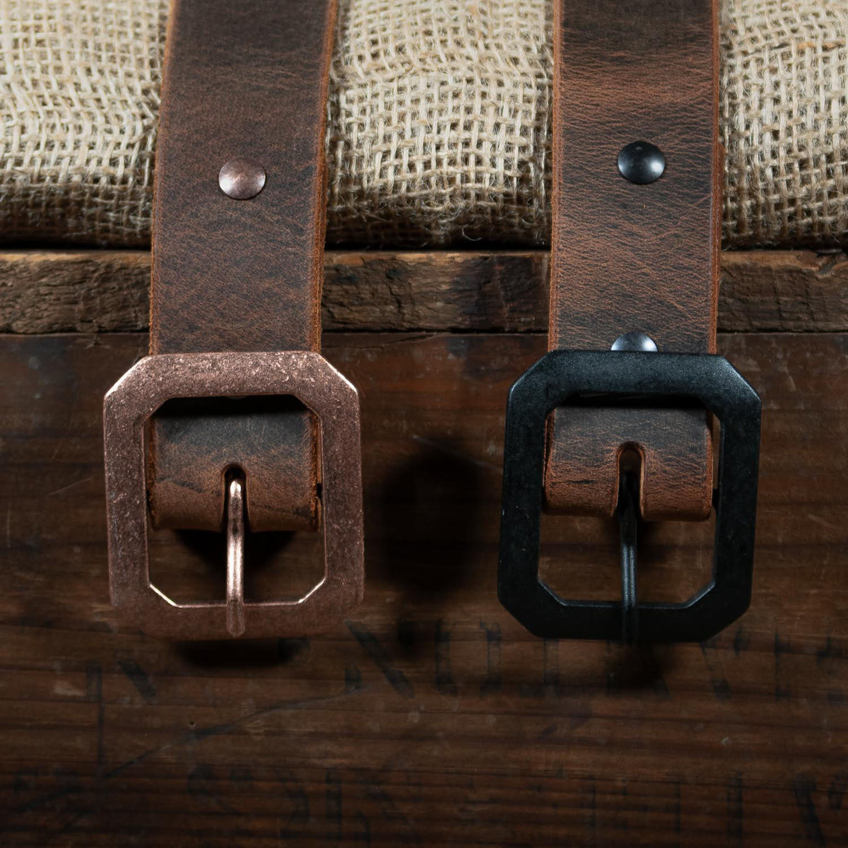 Two brown "The Relic Retro Style" belts by Hanks Belts, featuring rectangular copper and black buckles, rest side by side on wood with burlap behind them—showcasing classic American-made style.