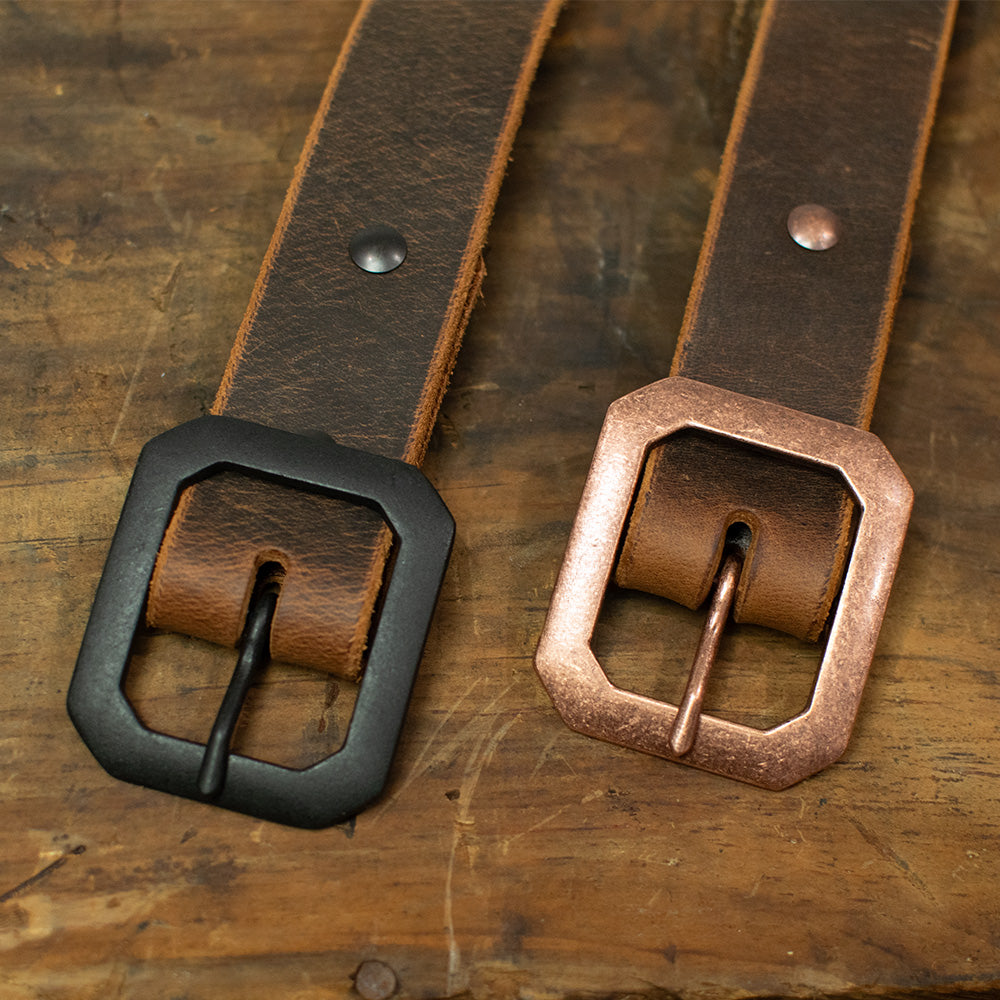 Two brown "The Relic Retro Style" belts by Hanks Belts rest side by side on wood—one with a matte black buckle, the other with a metallic copper buckle. Both square buckles have rounded corners, reflecting classic American-made style.