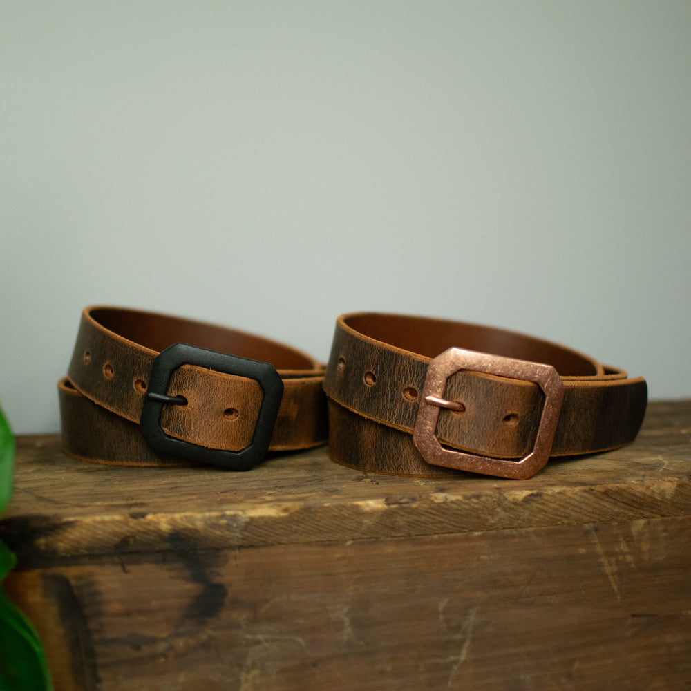 Two brown "The Relic Retro Style" belts by Hanks Belts, featuring black and copper square buckles, are shown on a rustic wooden surface with a light background. These American-made belts offer lasting durability and classic style.