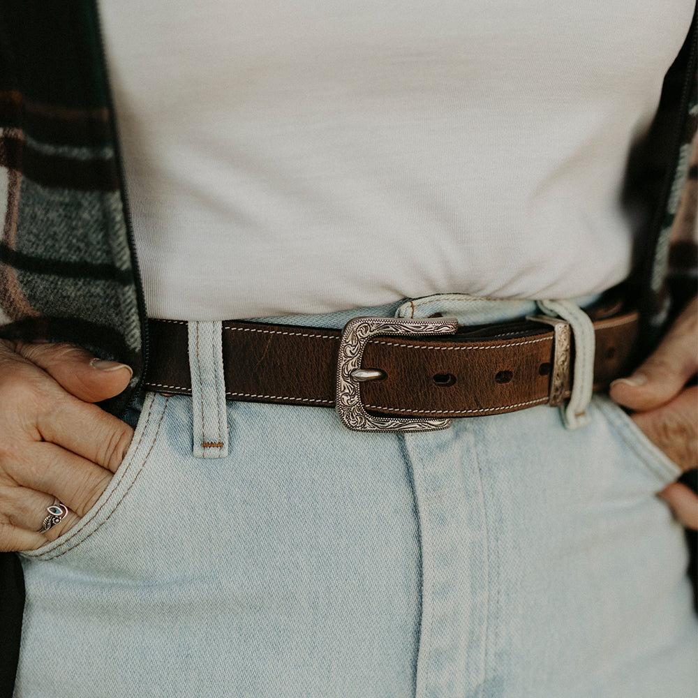 A person in light blue jeans, a white shirt, and a plaid layer stands with hands in pockets, wearing Hanks Belts' The Willow Bison women's western belt featuring an ornate floral buckle and durable backing for lasting style.