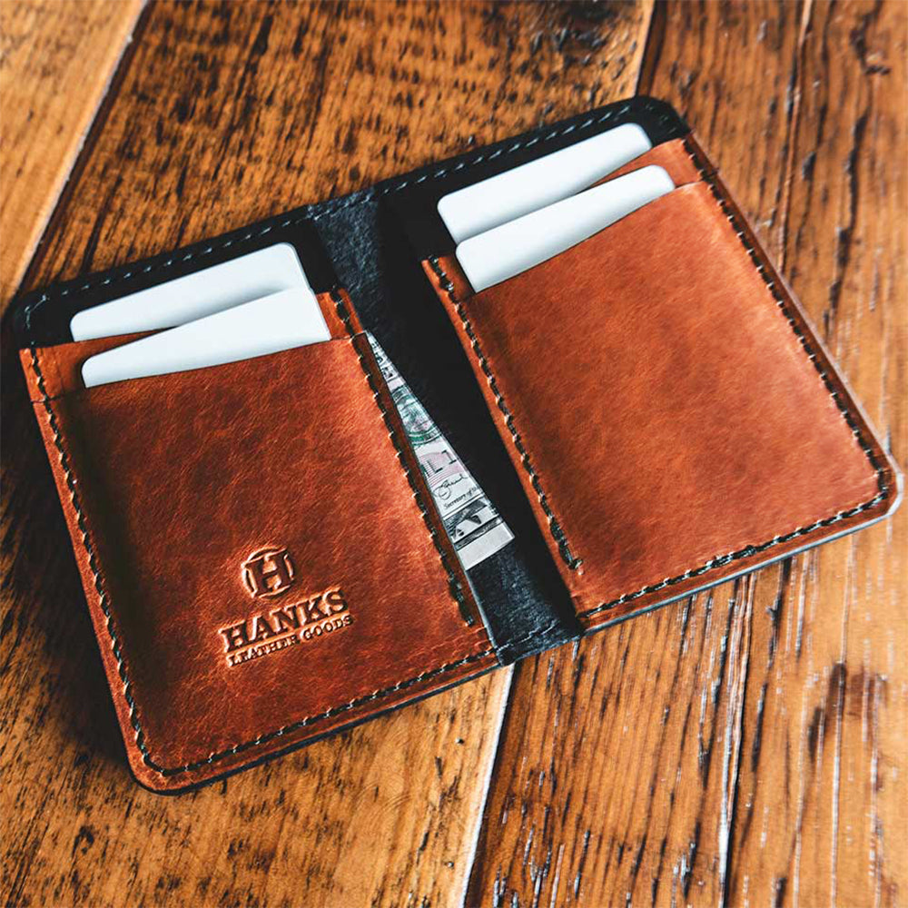 The Superior Front Pocket Bifold by Hanks Belts, crafted from premium brown harness leather, rests open on wood. It reveals two pockets with white cards and an embossed logo. A partially visible bill peeks out from one pocket.