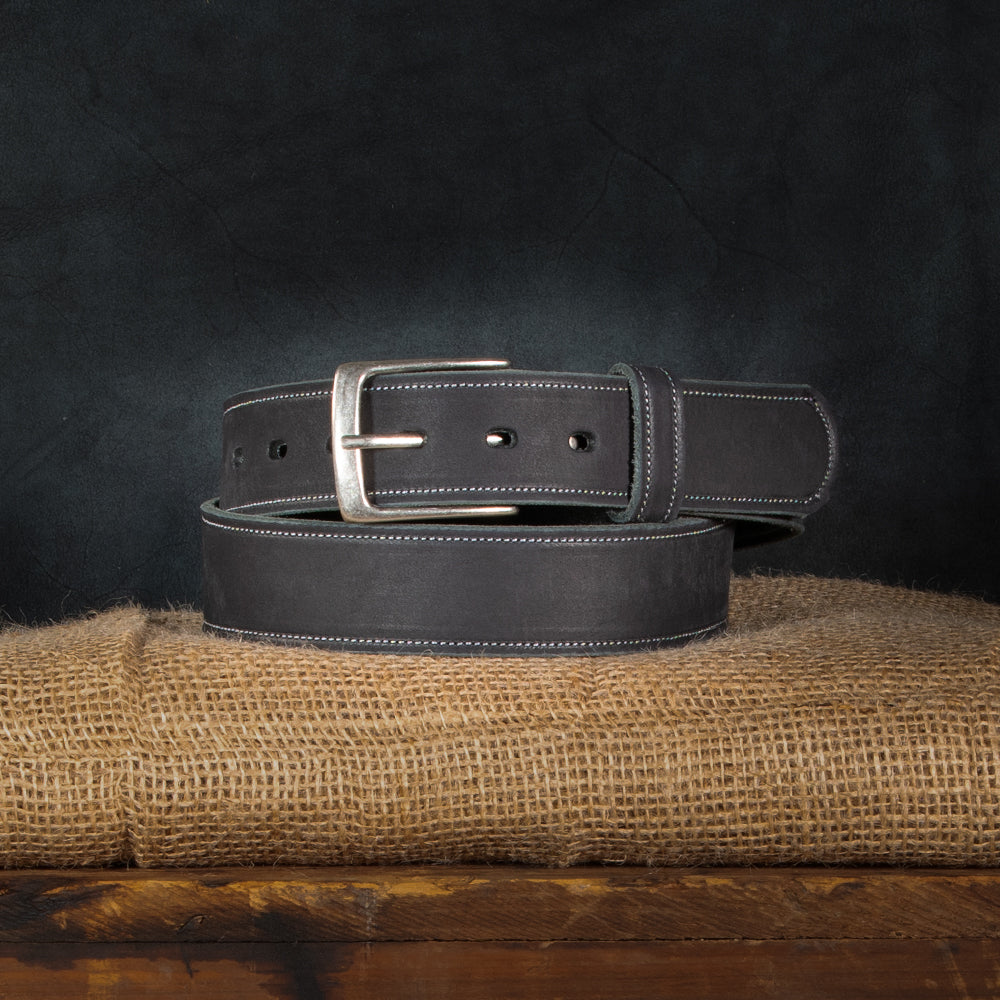 A neatly coiled Hanks Belts' USA-made "The Suede Belt - 1.5" in dark leather with a silver buckle sits on burlap, set against a dark, textured background that highlights its smooth finish and white stitching.