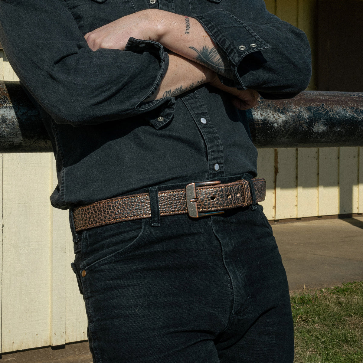 A person stands outdoors in front of a yellow building, arms crossed to display tattoos, wearing a dark denim shirt, black jeans, and The Yellowstone belt from Hanks Belts.