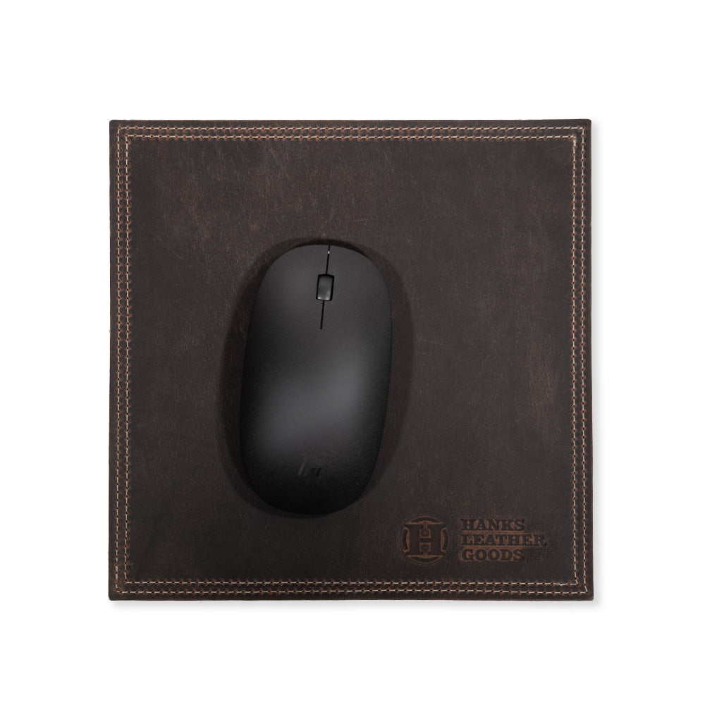 A black computer mouse rests on a dark brown bison leather mouse pad with decorative stitching, showcasing premium craftsmanship. This office accessory, branded by "Hanks Belts," features their logo embossed in the bottom right corner.