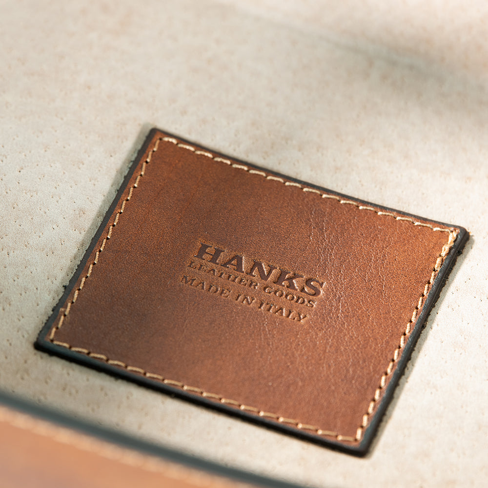 Close-up of a brown full-grain leather patch with stitching, engraved with the words "HANKS BELTS MADE IN ITALY." The background is a soft, neutral fabric, perfectly showcasing the travel-ready elegance of the Italian Large Valet Tray.