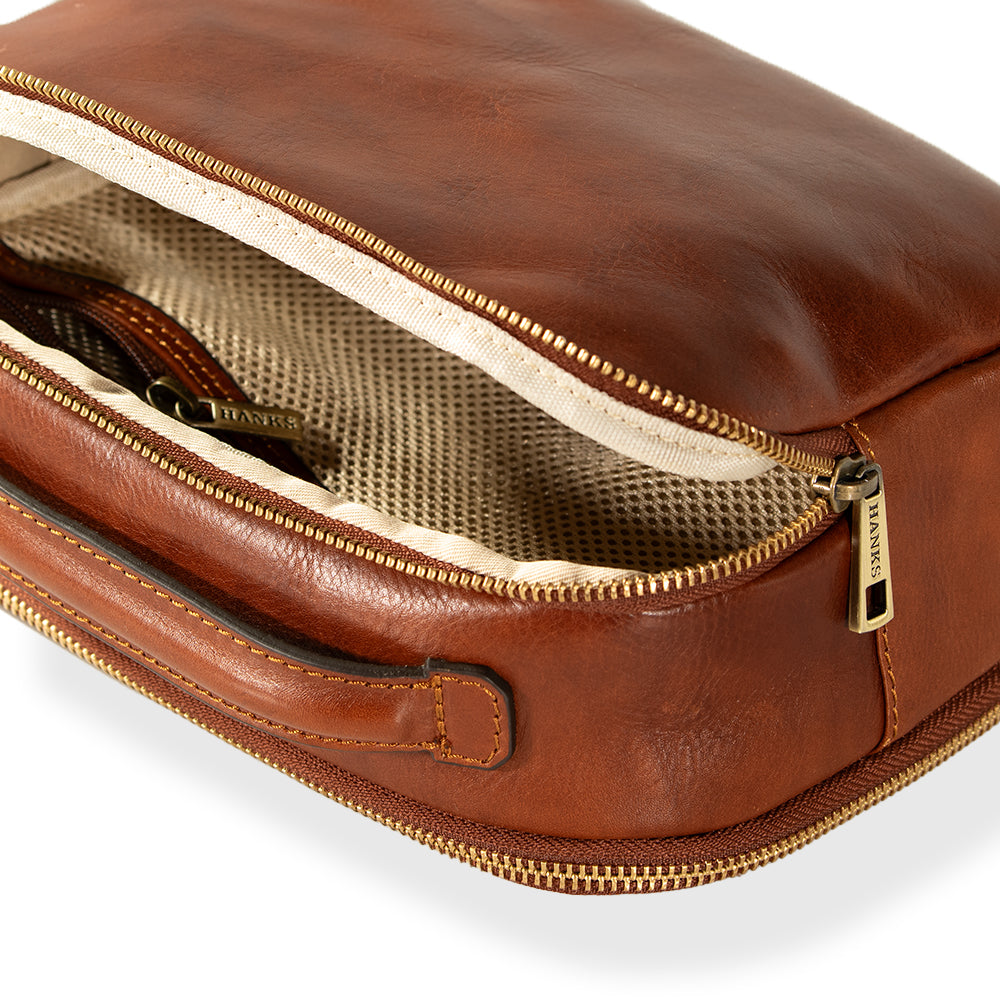Close-up of an open Tidy Travel Case by Hanks Belts, featuring a brown leather exterior with a visible zipper and a beige fabric interior. The gold zipper pull, engraved with "Hanks," showcases Italian craftsmanship. A leather handle is attached to the side.