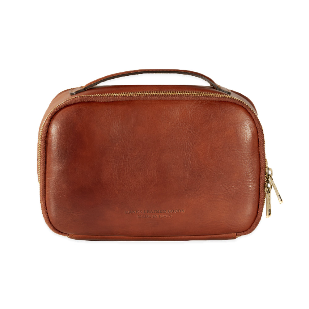 The Tidy Travel Case by Hanks Belts is crafted from brown Italian leather and features a handle on top with a zipper closure. Its smooth front has subtle text embossed near the bottom, providing an organized look. The dual pull tabs on the zipper make it an ideal travel companion.