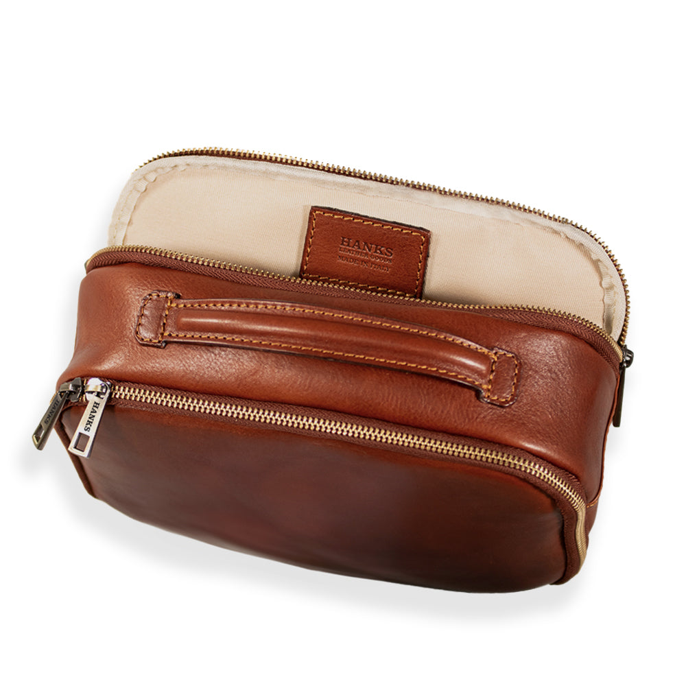 A luxurious Tidy Travel Case in brown leather with a beige interior, slightly open to showcase its contents. Highlighting exquisite Italian craftsmanship, this bag includes a zipper, a handle on top, and an inner label that reads "Hanks Belts." Perfect for organized travel.