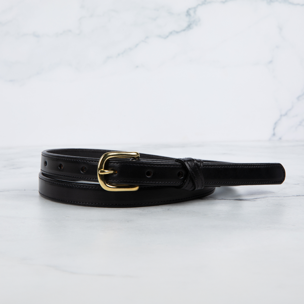 The Dixie by Hanks Belts, a black Premium English Bridle leather belt with a shiny gold buckle, is coiled on a white marble surface against a light gray marbled background.