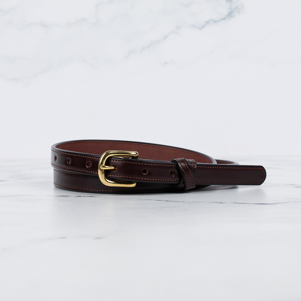 The Dixie by Hanks Belts—a dark brown women's belt made from premium English Bridle leather with a gold buckle—rests coiled on white marble against a light gray marbled background.