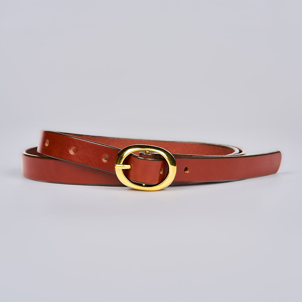 The Ivy by Hanks Belts is a slim Oak leather belt featuring a shiny gold oval buckle, coiled neatly against a plain light gray background.