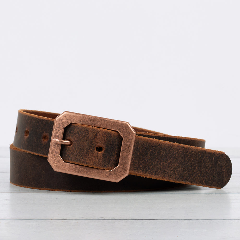 The Lainey by Hanks Belts is made from Full Grain Crazy Horse leather and showcases a large rectangular antique copper center bar buckle, coiled on a light wooden surface with a plain light gray background.