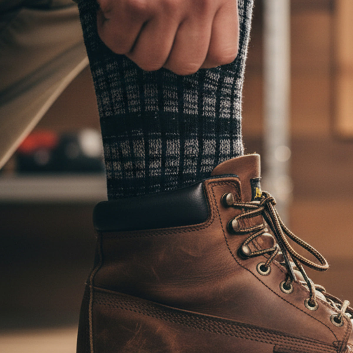 A close-up of a hand adjusting a Hank Belts Upland Sock in black and white plaid above a brown leather hiking boot with metal eyelets and laces, set against a softly blurred background.