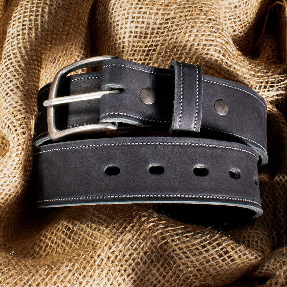 Elegant close-up of Hanks Belts' USA-made "The Suede Belt - 1.5"" in black, featuring a silver buckle, white stitching, and multiple holes. The belt is gracefully coiled on burlap fabric with sunlight casting shadows across the scene.