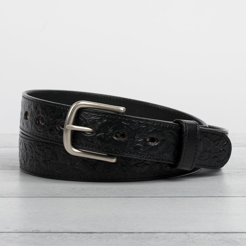 The Floral Embossed by Hanks Belts, a black full-grain leather belt with a silver buckle and floral embossing, is coiled on a light gray wooden surface against a plain white background.