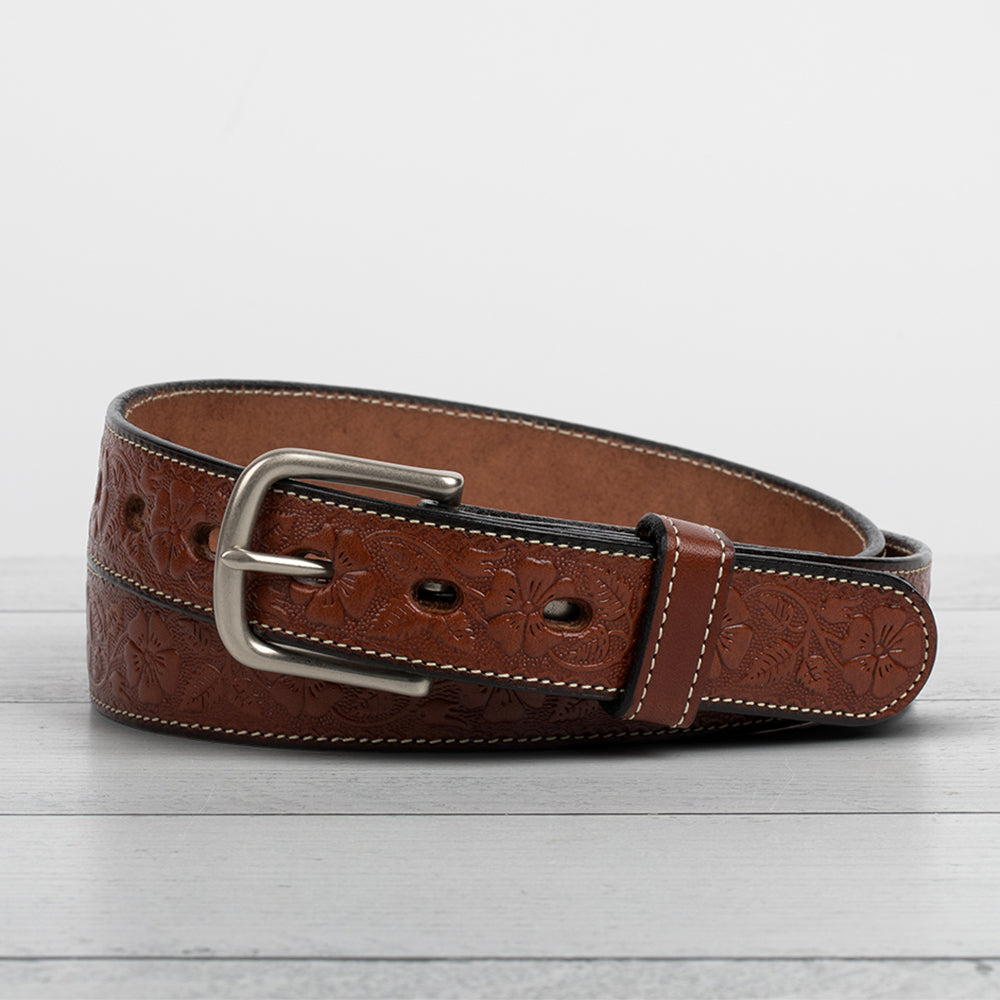 The Floral Embossed by Hanks Belts, a full-grain leather belt with floral embossing, light stitching, and a silver buckle, is coiled on a light gray wooden surface.