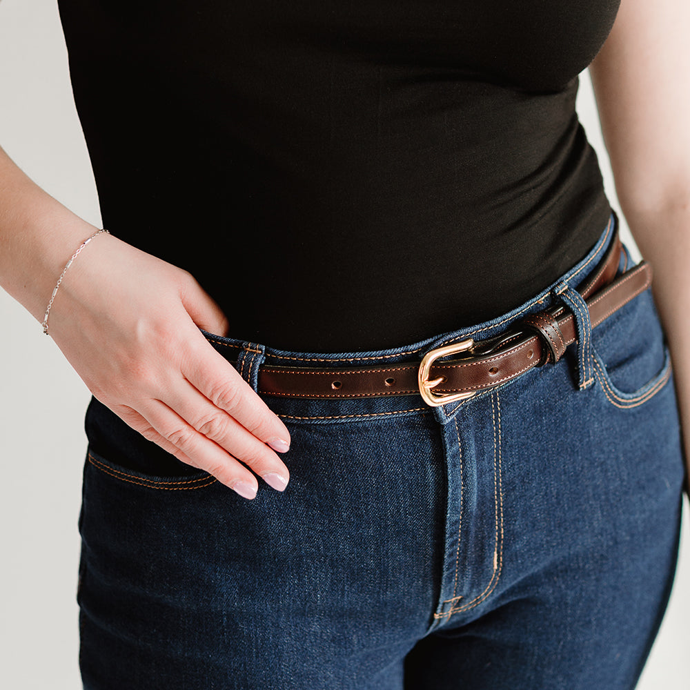 A person in a black top and blue jeans poses with manicured nails and a delicate bracelet, showcasing Hanks Belts' The Dixie women's belt—crafted from premium English Bridle leather with a gold buckle.