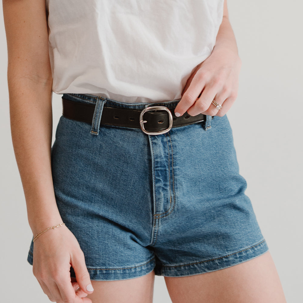 A person wears a white t-shirt tucked into high-waisted blue denim shorts with The Eloise by Hanks Belts. Their left hand, adorned with a ring and bracelet, rests near the waistband. The background is plain and light-colored.
