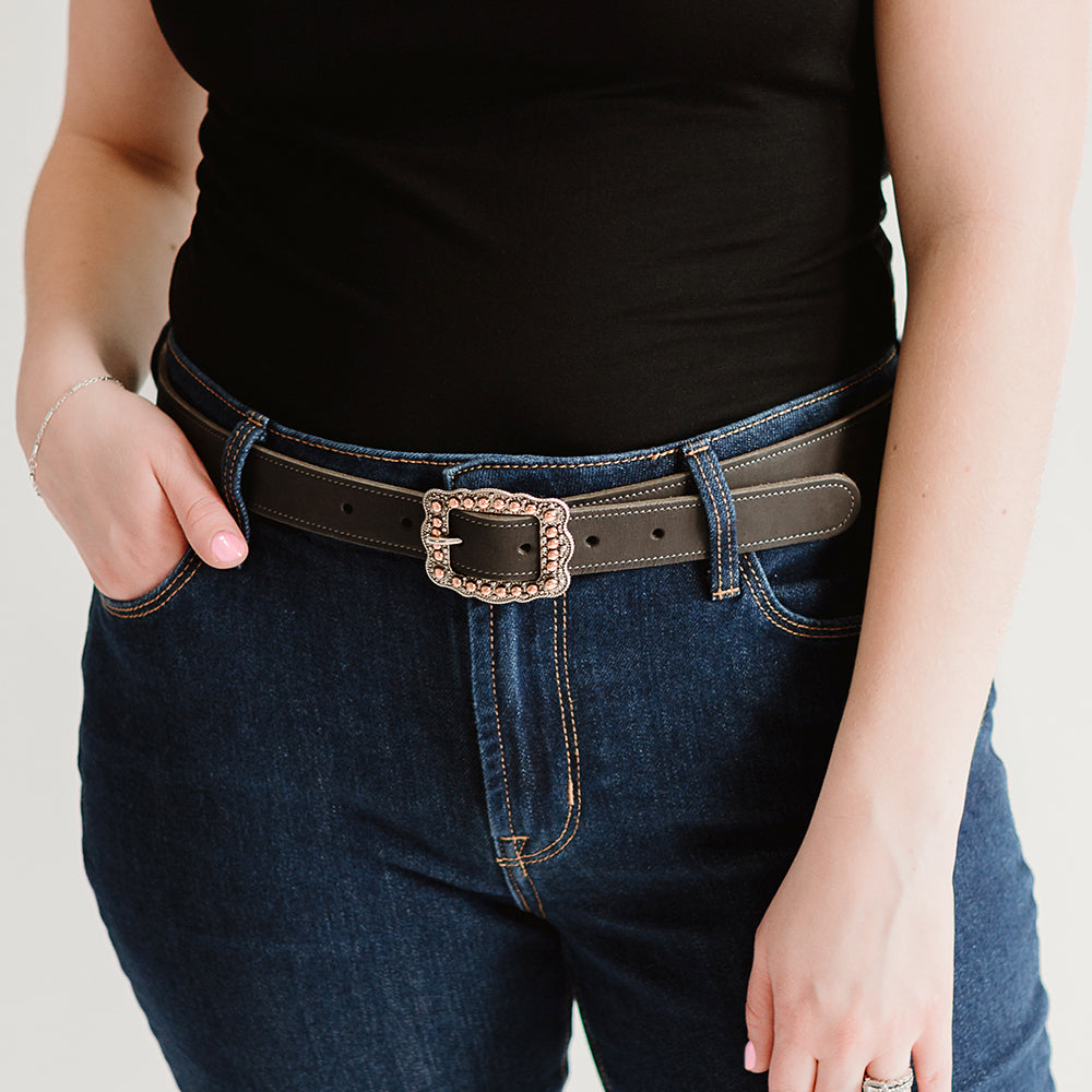 A person in a black shirt and dark blue jeans wears The Lennon Suede by Hanks Belts, featuring a decorative rectangular rose gold buckle, with one hand in their pocket.