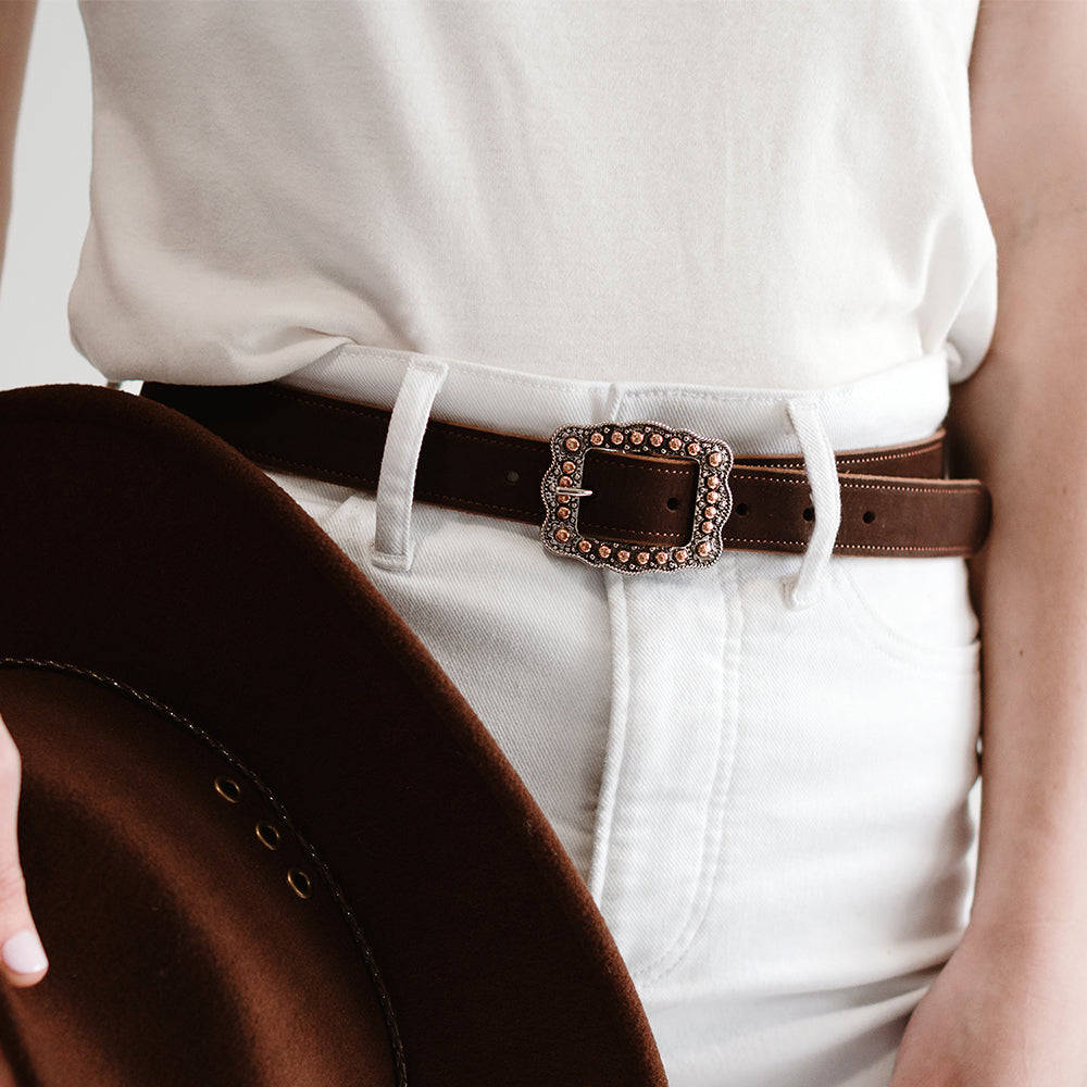A person wears a white top tucked into high-waisted white jeans, accessorized with The Lennon Suede brown Western belt by Hanks Belts featuring a rose gold buckle, and holds a brown wide-brimmed hat at their side.