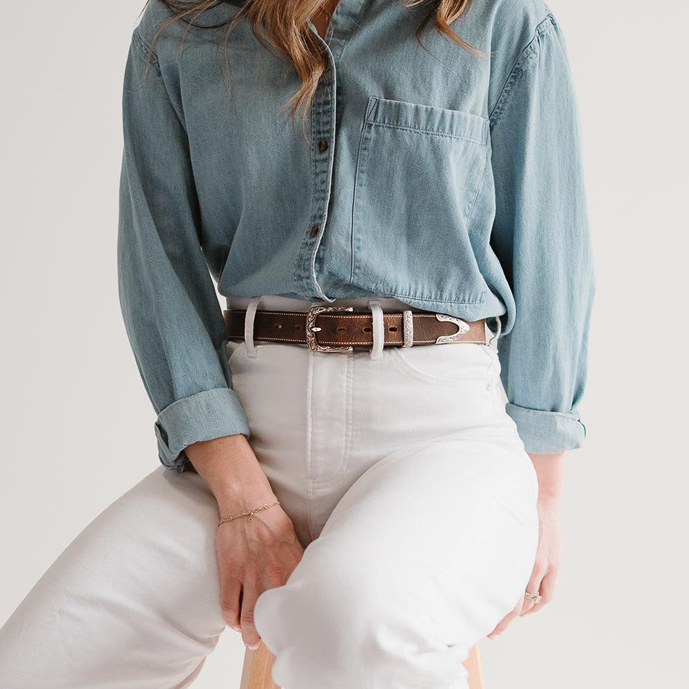 A person sits on a wooden stool, wearing a light blue button-up shirt tucked into high-waisted white pants, styled with Hanks Belts’ The Willow Bison women's western belt featuring a floral buckle. Their face is not visible.