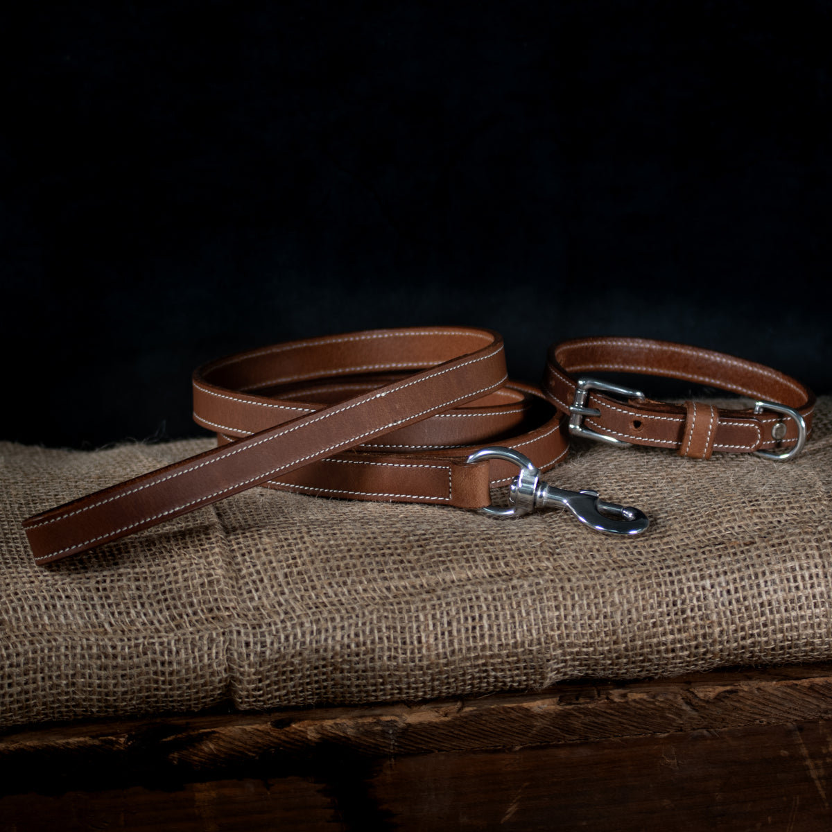 The Ollie Bison Collar by Hanks Belts, made from premium brown bison leather with white stitching and stainless steel hardware, is displayed on burlap against a dark background.