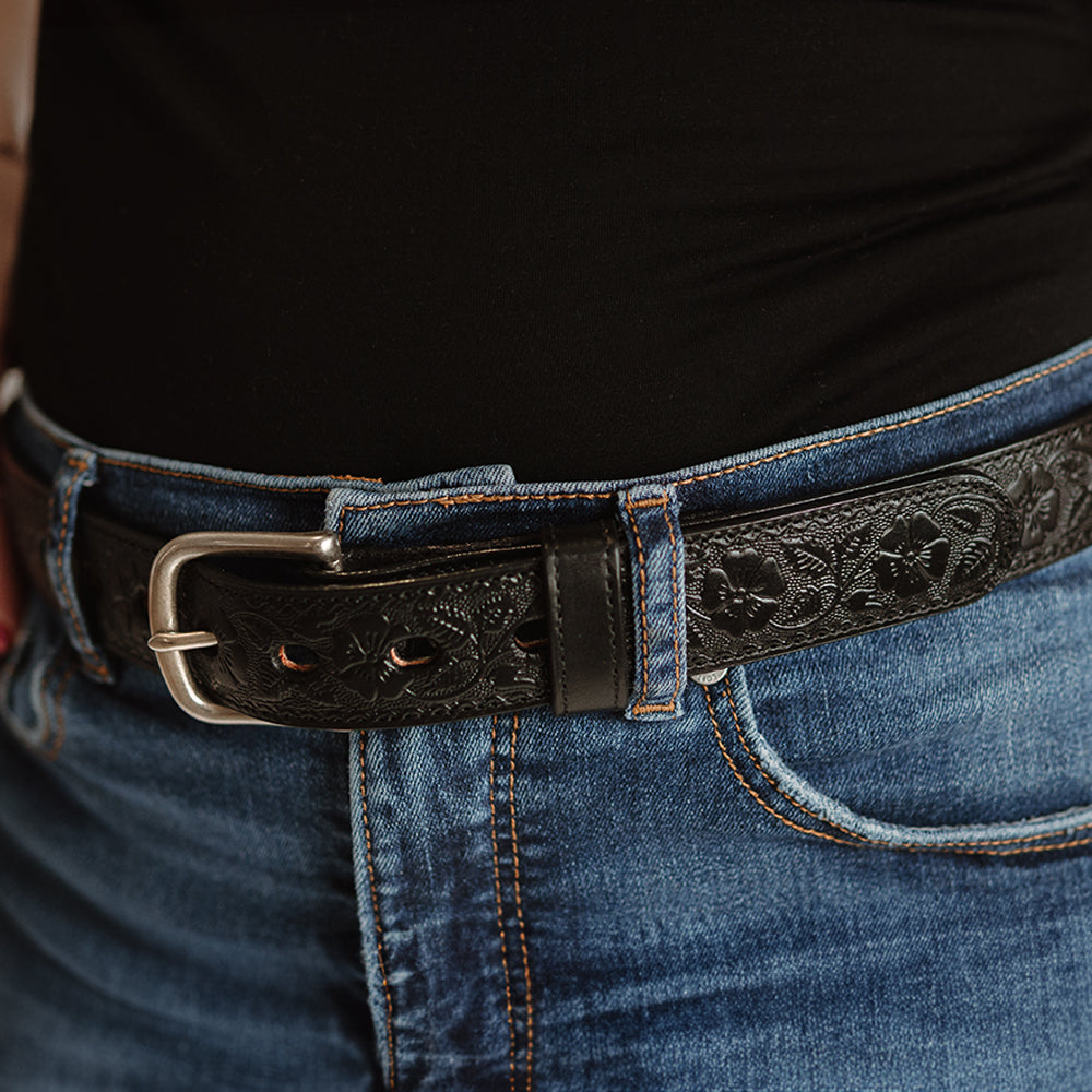 Close-up of a person wearing blue jeans and a black shirt, featuring The Floral Embossed by Hanks Belts—a full-grain leather, USA-made belt with an embossed floral design and silver buckle.
