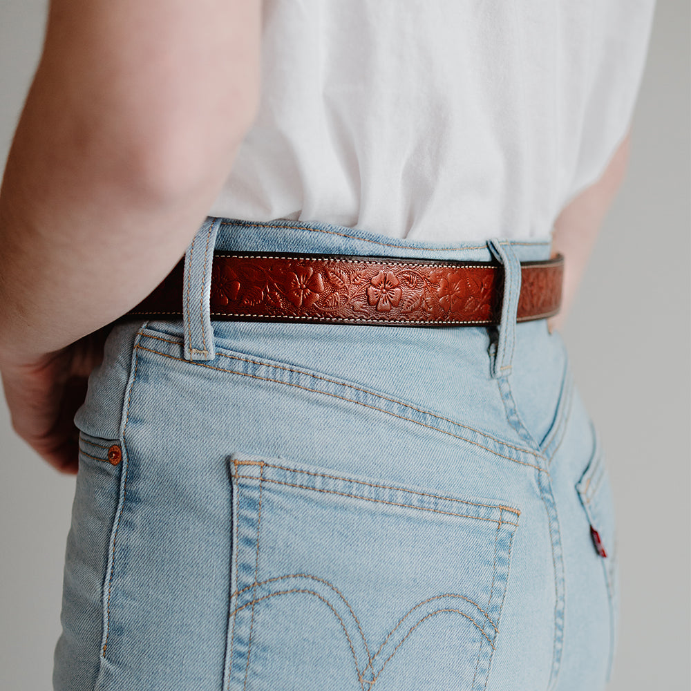 A person wearing light blue jeans and a white shirt, shown from the waist down, with Hanks Belts' The Floral Embossed—a full-grain leather belt—fastened around their jeans.