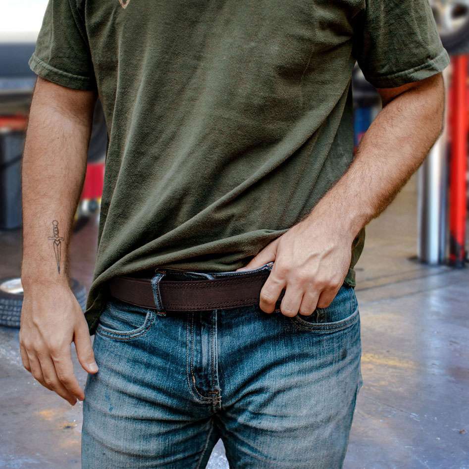 A person in a green t-shirt and blue jeans stands indoors, adjusting their Hanks Belts "The Scratchless Mechanics" belt. Their tattooed left arm is visible, with vehicle tires and equipment blurred in the background.