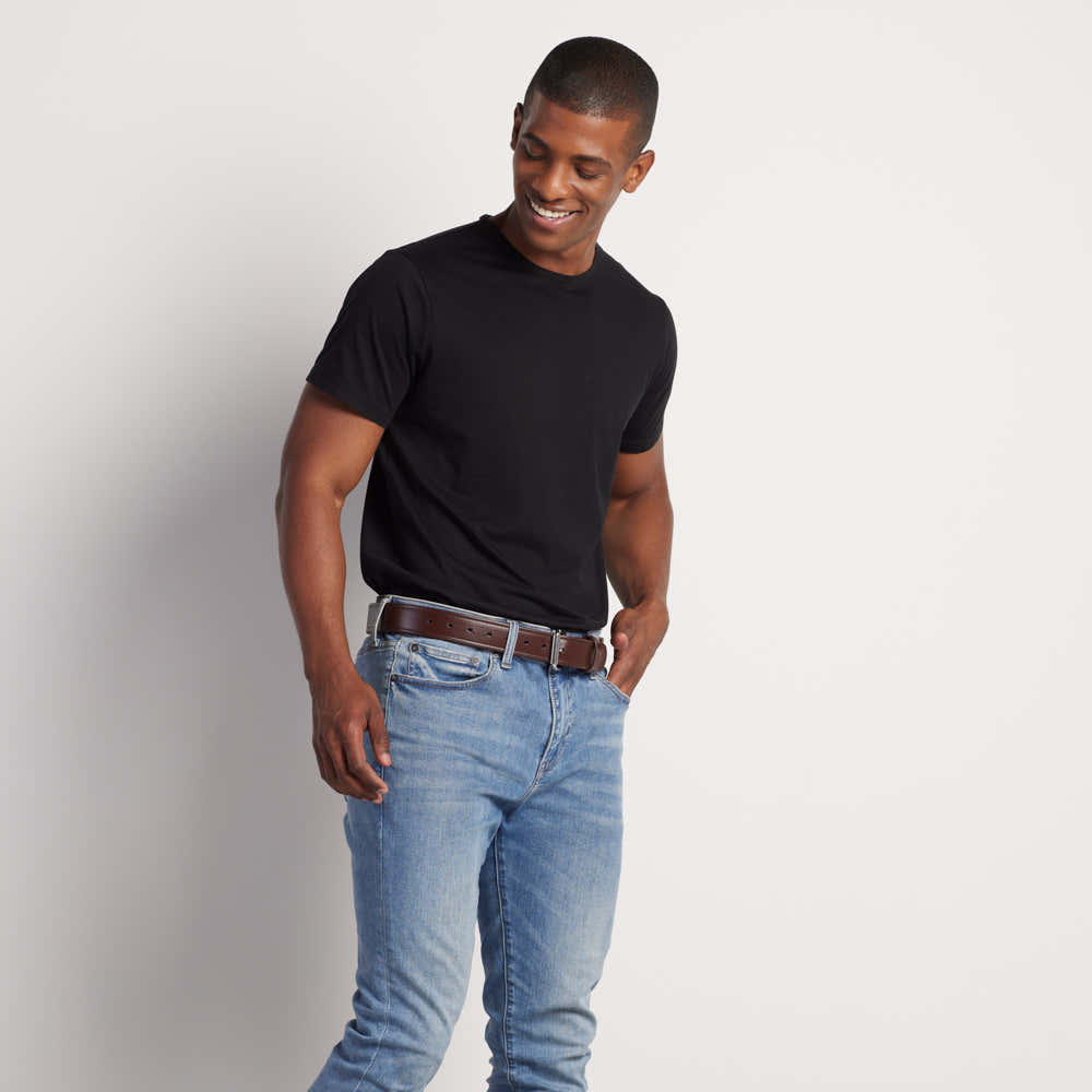 A man in a black t-shirt and light blue jeans stands against a white background, smiling with one hand in his pocket while showcasing Hanks Belts’ The Sweat & Waterproof Belt Synthetic Double Layer.