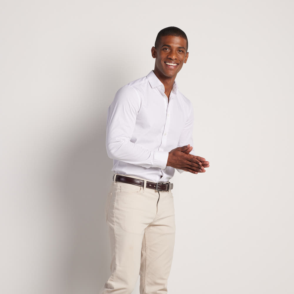 A smiling man stands against a white background, wearing a white dress shirt tucked into beige pants with Hanks Belts' The Esquire Raised, crafted from English Bridle leather. His hands are loosely clasped in front of him.