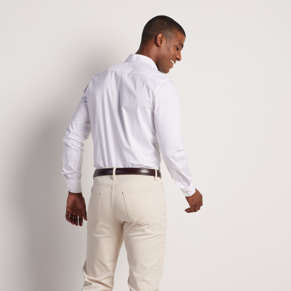 A man stands smiling, facing away from the camera in a white button-up shirt and cream pants, wearing The Esquire Raised belt by Hanks Belts, crafted from English Bridle leather, against a plain light background.