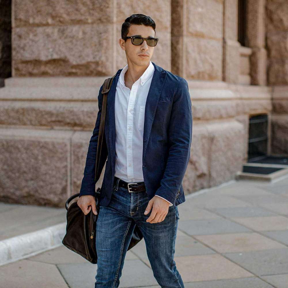 A man in sunglasses, a navy blazer, white shirt, and jeans walks by a stone building with a dark leather bag, wearing Hanks Belts' The Esquire Raised belt crafted from English Bridle leather.