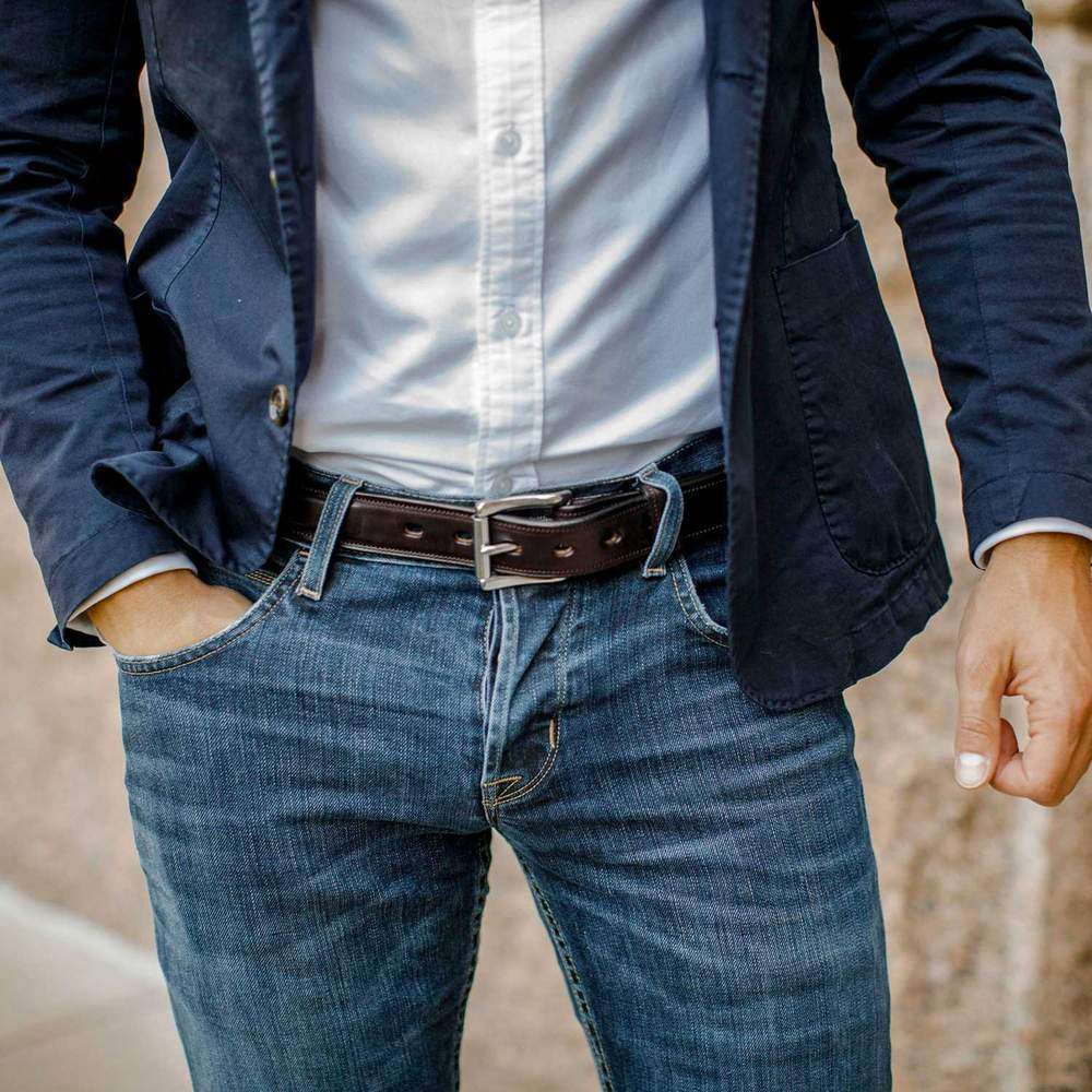 A person wears a navy blazer, white button-up, blue jeans, and The Esquire Raised belt by Hanks Belts. One hand is in their pocket. The photo is cropped to show only the torso and upper legs.