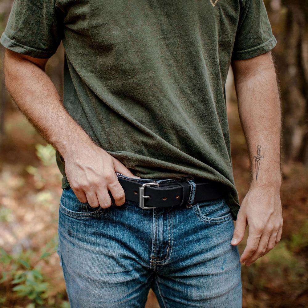 A person in a green t-shirt and blue jeans adjusts a black Hanks Belts The Gunner belt, made from full-grain steer hide. A dagger tattoo is visible on their left forearm, with blurred trees in the outdoor background.