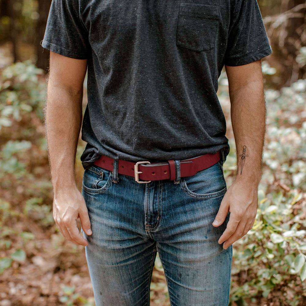 Someone stands outdoors in a wooded area, wearing a dark T-shirt, blue jeans, and Hanks Belts' The Gunner belt in red. Only their torso and upper legs are visible, with hands relaxed at their sides.