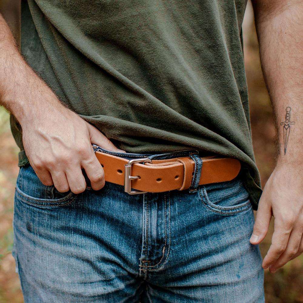 A person in a green t-shirt and blue jeans adjusts The Gunner belt from Hanks Belts, crafted from full-grain steer hide. Their partially visible left arm features a dagger tattoo on the inner forearm.