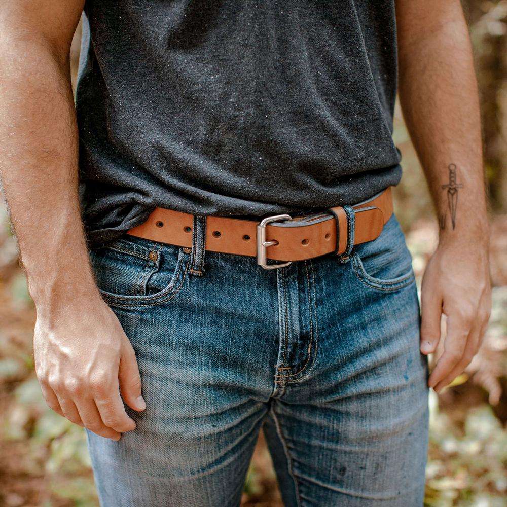 Outdoors, a person in a black t-shirt, blue jeans, and The Gunner full-grain leather belt by Hanks Belts stands with relaxed hands at their sides. A sword tattoo is visible on their left forearm.