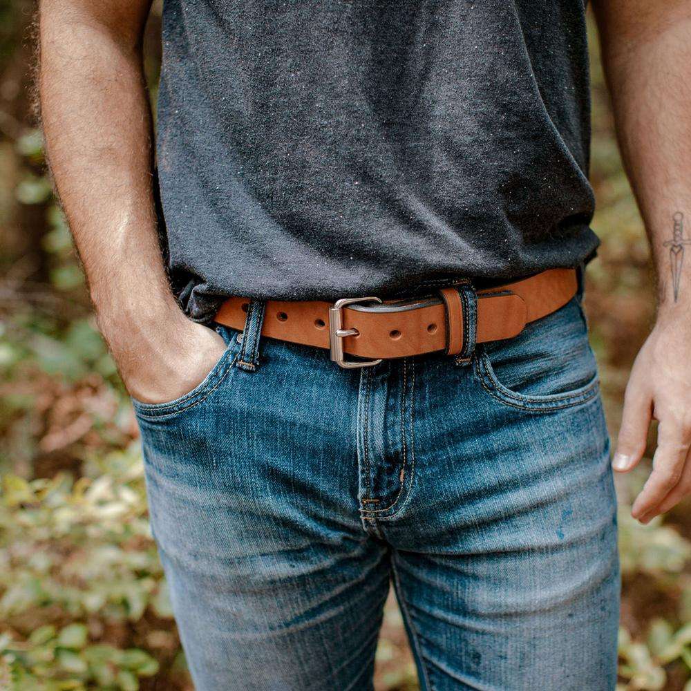 A person wearing a dark grey t-shirt, blue jeans, and The Gunner by Hanks Belts stands outdoors with one hand in their pocket. Only the torso and upper legs are visible.