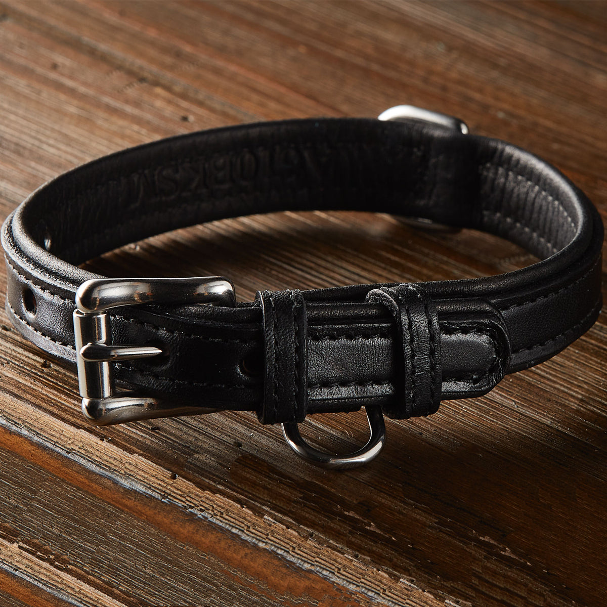 A close-up view of the Rover Standard Collar by Hanks Belts, a USA Made premium leather collar in black with a silver metal buckle and D-ring, placed on a wooden surface. The collar features stainless steel hardware, multiple size adjustment holes, and a sleek, durable design.