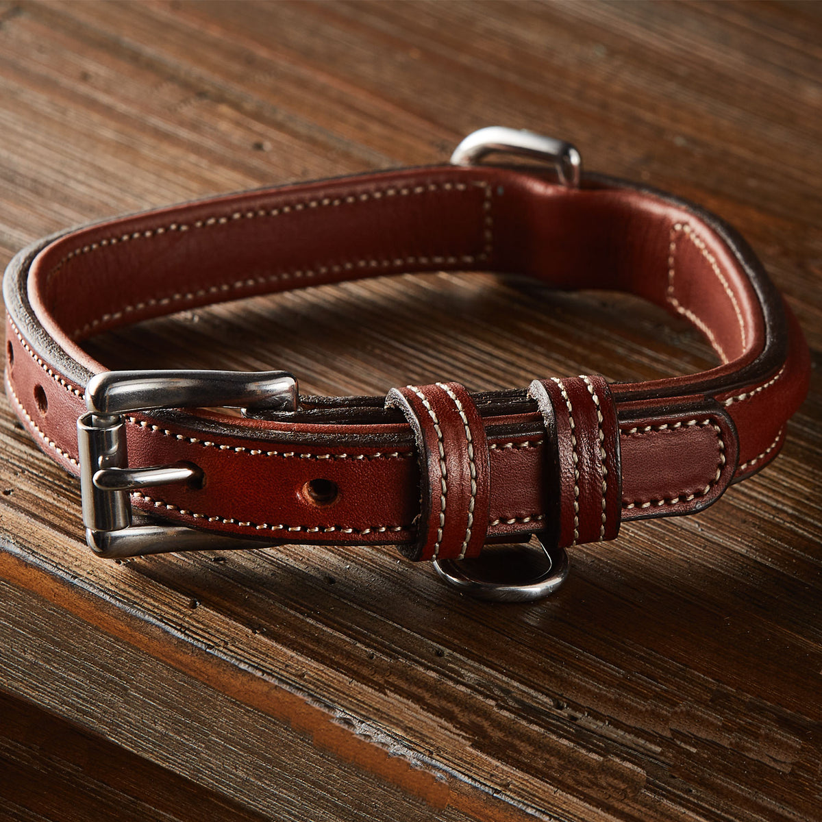 The Bentley Raised Collar by Hanks Belts is a brown English bridle leather dog collar with white stitching and a stainless steel buckle, displayed on a wooden surface.